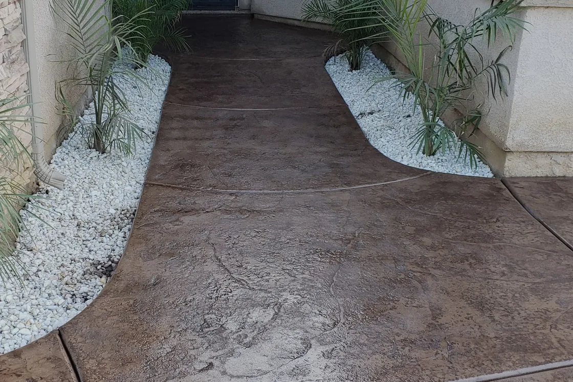 Finished stamped concrete walkway curving between white rock beds and palms