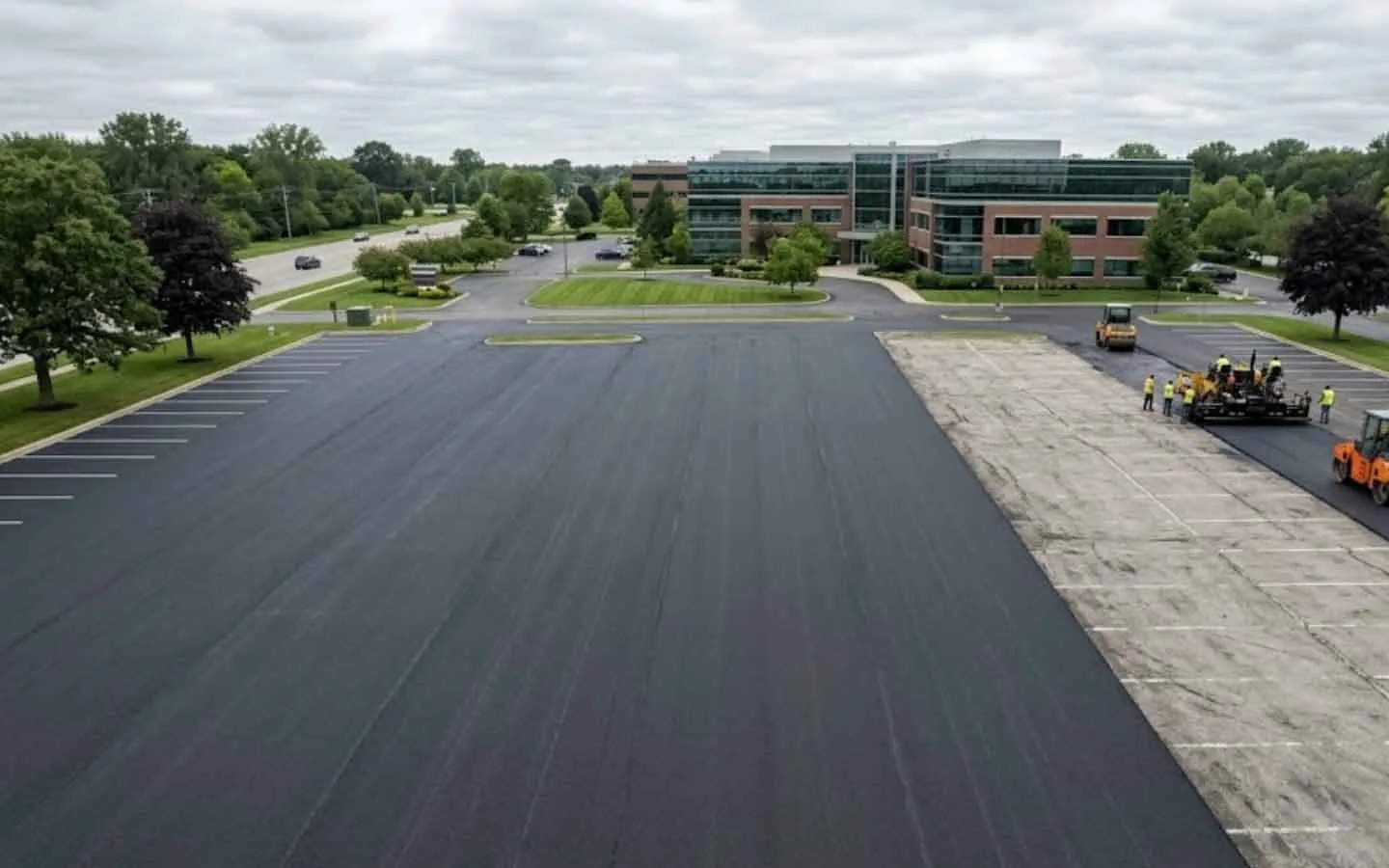 Half of a parking lot overlaid with new asphalt while the crew paves the rest, asphalt resurfacing