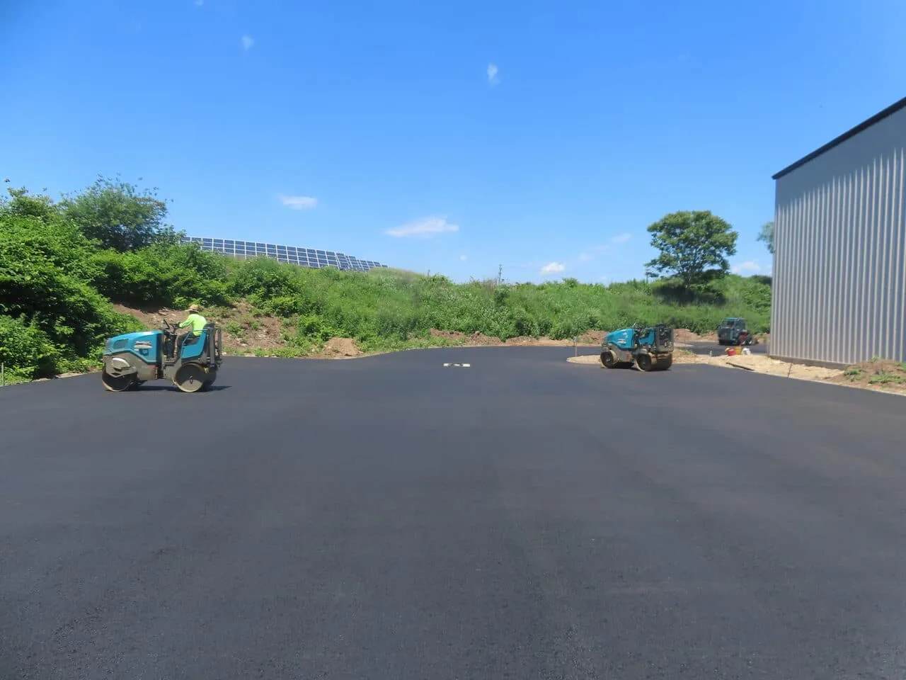 Two rollers compacting a new asphalt parking lot beside a metal building, commercial paving