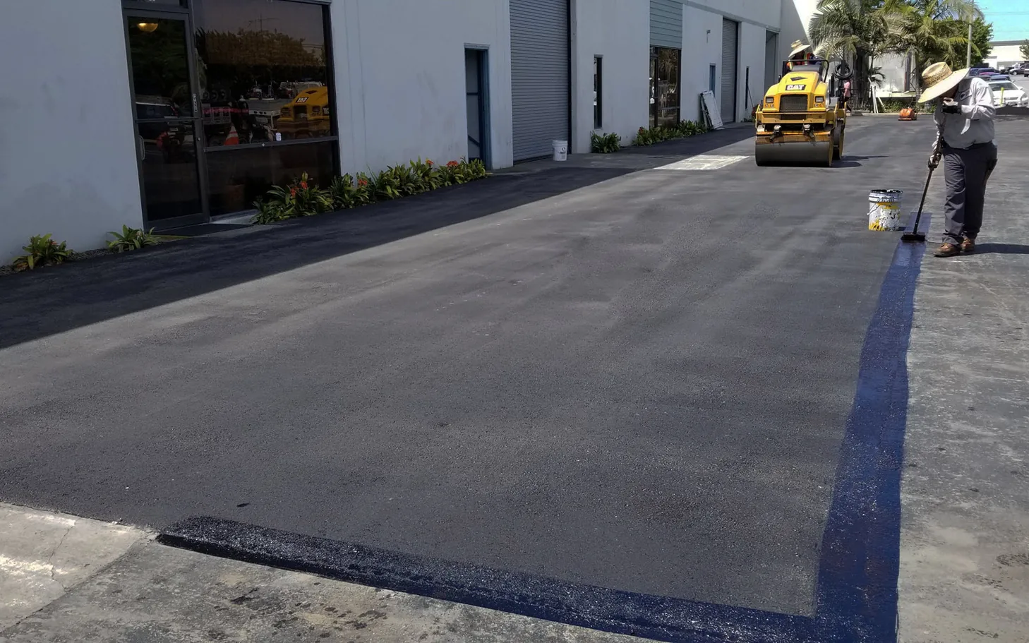 Worker painting a blue stall line on new asphalt as a roller finishes the lot, parking lot striping